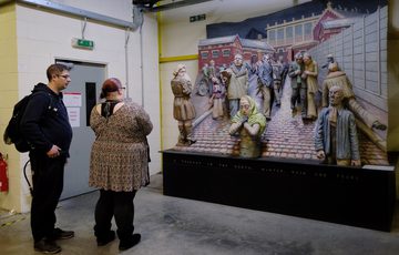 Two adults looking at a large 3D artwork which shows people in a city street.