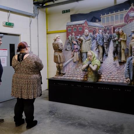 Two adults looking at a large 3D artwork which shows people in a city street.