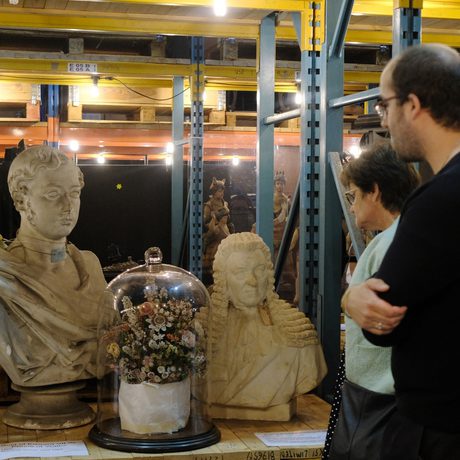 Two adults looking at two busts and a terrarium containing flowers that are on open display on storage shelving.