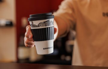 Hand holding a black and white travel coffee cup labeled "Birmingham" with city skyline.