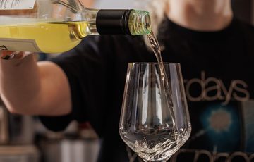A person with glasses pours white wine into a wine glass in a cozy setting.
