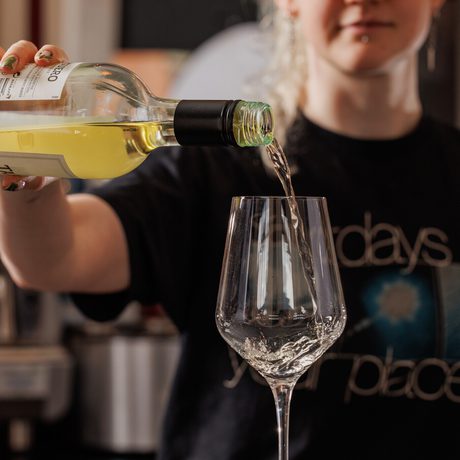 A person with glasses pours white wine into a wine glass in a cozy setting.