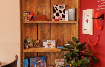 Wooden shelves hold board games next to a red wall with feedback phone.
