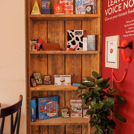 Wooden shelves hold board games next to a red wall with feedback phone.