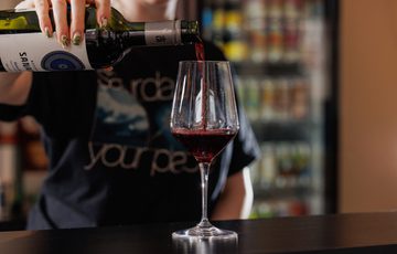 A person pours red wine from a bottle into a wine glass on a counter.