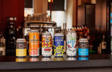 Six assorted craft beer cans lined up on a bar counter with a blurred background.