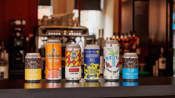 Six assorted craft beer cans lined up on a bar counter with a blurred background.