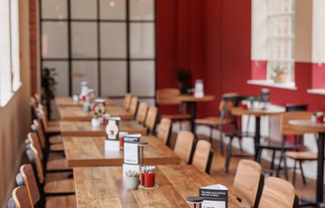 Long wooden tables and chairs line a bright, modern cafe bar area with red walls and hanging lights.