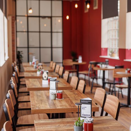 Long wooden tables and chairs line a bright, modern cafe bar area with red walls and hanging lights.