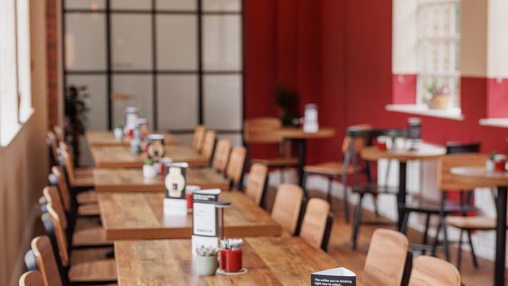 Long wooden tables and chairs line a bright, modern cafe bar area with red walls and hanging lights.