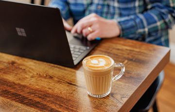 Clear glass of latte with latte art on wooden table, person working on laptop nearby.