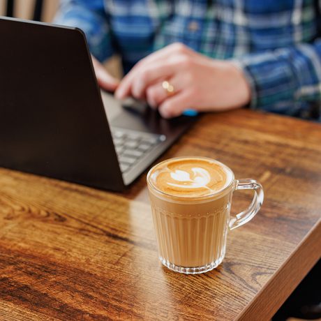 Clear glass of latte with latte art on wooden table, person working on laptop nearby.