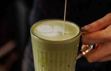 Pouring milk into a glass of matcha latte with latte art in progress.