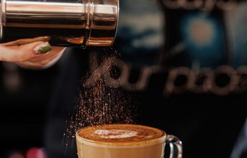A hand sprinkles cocoa or cinnamon powder onto a latte in a clear glass mug.