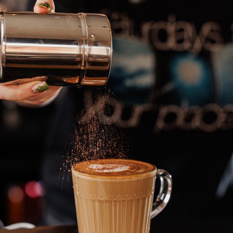 A hand sprinkles cocoa or cinnamon powder onto a latte in a clear glass mug.