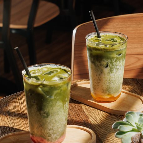Two iced matcha drinks with layered colours sit on a wooden table in sunlight.