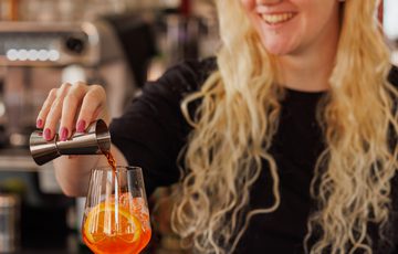 A smiling woman with glasses pours a drink into a wine glass at a bar.