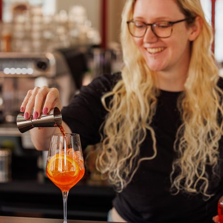 A smiling woman with glasses pours a drink into a wine glass at a bar.