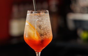 A refreshing orange cocktail with ice and a straw in a wine glass on a dark worktop.