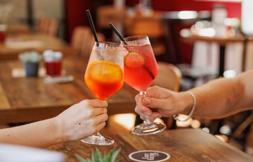 Two people's hands clinking colourful cocktails in a cozy café setting.