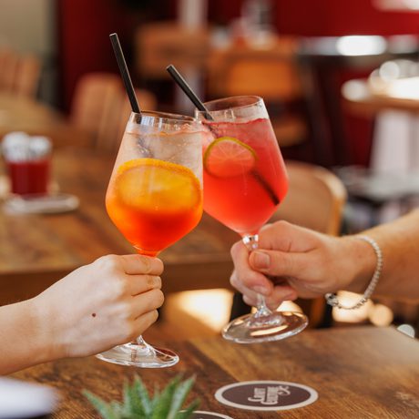 Two people's hands clinking colourful cocktails in a cozy café setting.