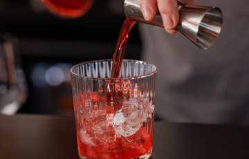 Man pours liquid from jigger into glass with ice, preparing a red cocktail.