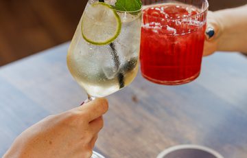 Two people clinking colourful cocktails with straws over a wooden table in a cozy setting.