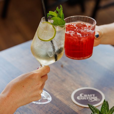 Two people clinking colourful cocktails with straws over a wooden table in a cozy setting.