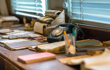 Vintage rotary phone, scattered papers, notebooks, and pencils on a cluttered wooden desk.