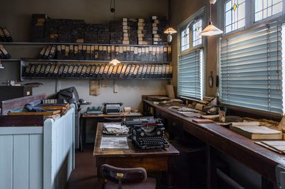 An historic office with large wooden desk by the window, typewriter and folders stored on three shelves on a wall.