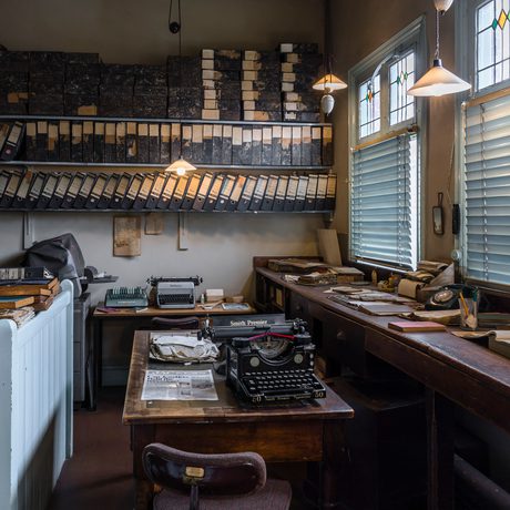 An historic office with large wooden desk by the window, typewriter and folders stored on three shelves on a wall.