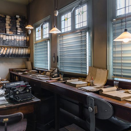 Vintage office with typewriters, paperwork, window blinds, wooden desks, and hanging lights.
