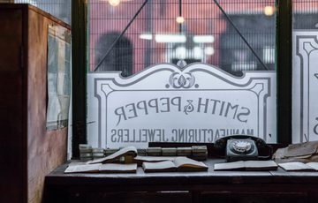 Desk with notebooks and a dial telephone in front of a window etched with a sign for 'Smith and Pepper'