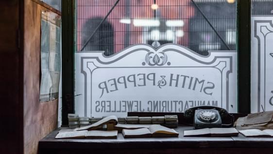 Desk with notebooks and a dial telephone in front of a window etched with a sign for 'Smith and Pepper'