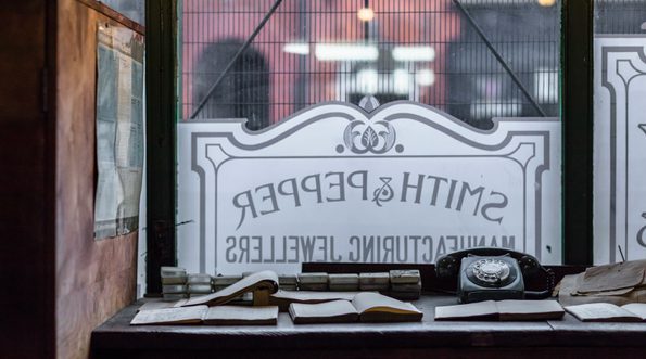 Desk with notebooks and a dial telephone in front of a window etched with a sign for 'Smith and Pepper'