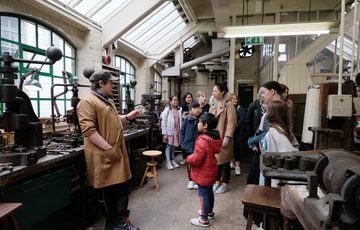 A staff member giving a tour to a group of adults and children in an historic jewellery workshop.