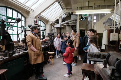 A staff member giving a tour to a group of adults and children in an historic jewellery workshop.