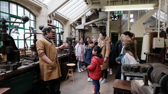 A staff member giving a tour to a group of adults and children in an historic jewellery workshop.