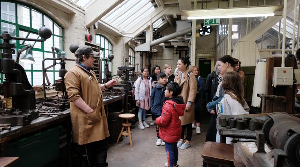 A staff member giving a tour to a group of adults and children in an historic jewellery workshop.