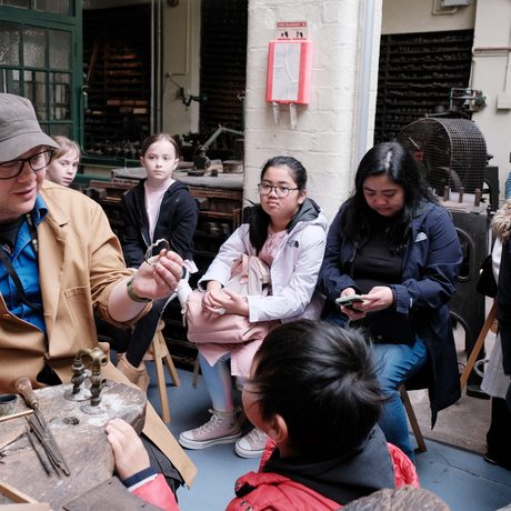 A group of children looking a staff member who is sitting at a workbench and holding an item of jewellery.