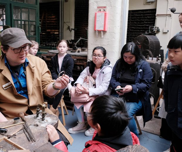A group of children looking a staff member who is sitting at a workbench and holding an item of jewellery.
