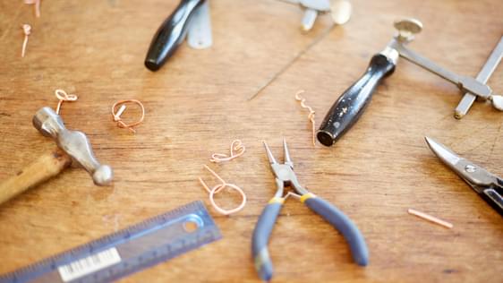 Jewellery making tools and metal in the process of becoming rings, laid out on a wooden table.