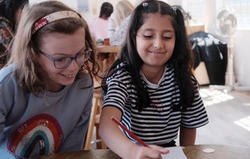 Two children smiling. They are sitting at a table, one child is writing on paper while the other looks on.