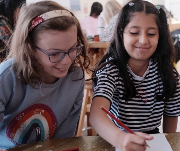 Two children smiling. They are sitting at a table, one child is writing on paper while the other looks on.
