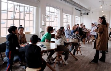 A group of adults and children sitting at tables listening to members of staff talking.
