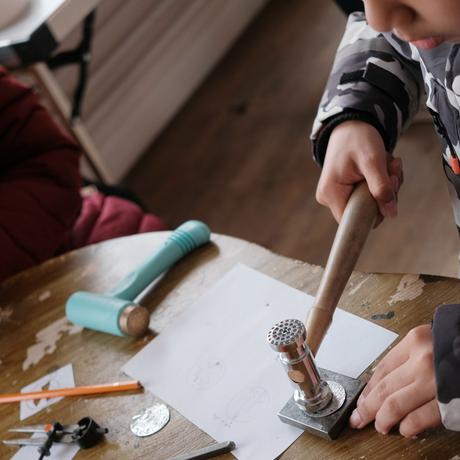 A child holding a jewellery making hammer and pressing it against a flat silver circle that is on a table.