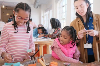 A member of staff with two smiling children. Jewellery making tools are on the table in front of them.
