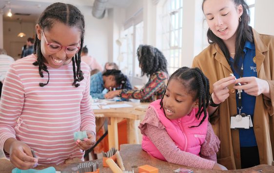 A member of staff with two smiling children. Jewellery making tools are on the table in front of them.