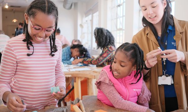 A member of staff with two smiling children. Jewellery making tools are on the table in front of them.