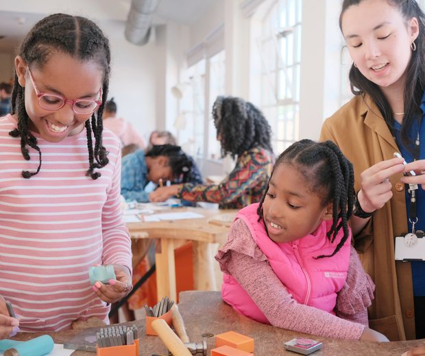 A member of staff with two smiling children. Jewellery making tools are on the table in front of them.